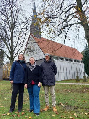 Drei Menschen vor einem Kirchgebäude, das zum Teil von Planen bedeckt ist.